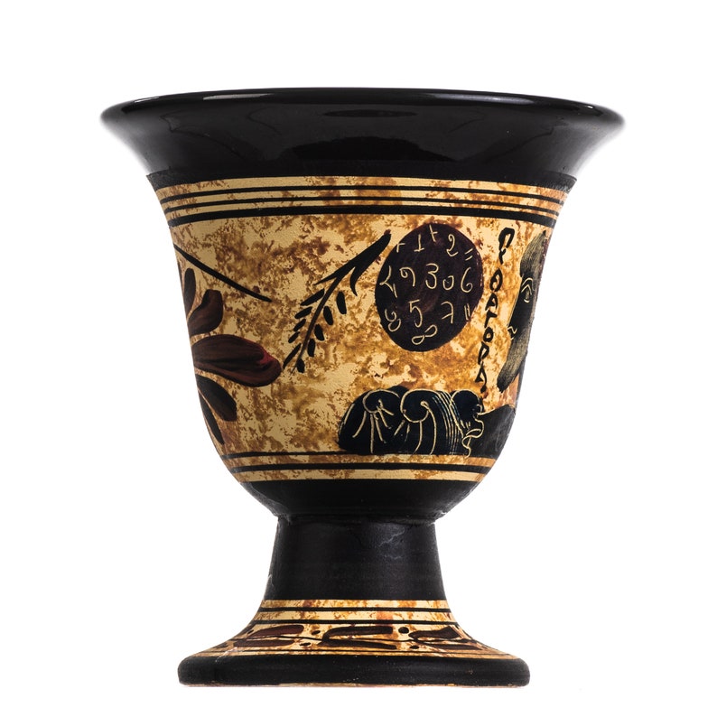 May include: A black ceramic goblet with a decorative design in shades of brown, gold, and black. The design features a stylized floral motif, a circular symbol with Greek letters, and a figure with a beard. The goblet has a wide, round base.