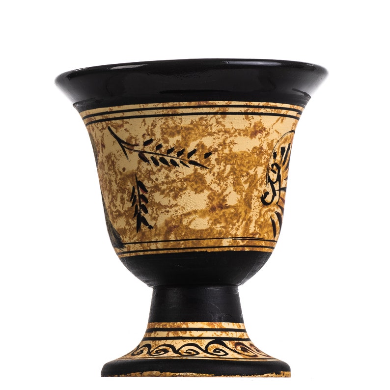 May include: A black and gold ceramic chalice with a pedestal base. The chalice features a black rim and base, with a gold-colored body adorned with black leaf and scroll designs. The design is reminiscent of ancient Greek pottery.