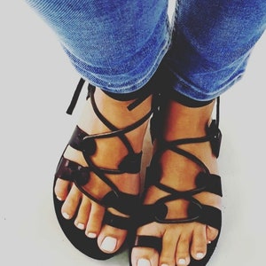 May include: Black leather sandals with lace-up straps and a buckle closure. The sandals have a flat sole and are worn with blue jeans.