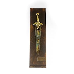 May include: A gold-colored dagger with a green patina is mounted on a dark wood plaque. The plaque has a brass plate with the text "Bronze Age Dagger, 1700-1500 BC, National Museum of Denmark".