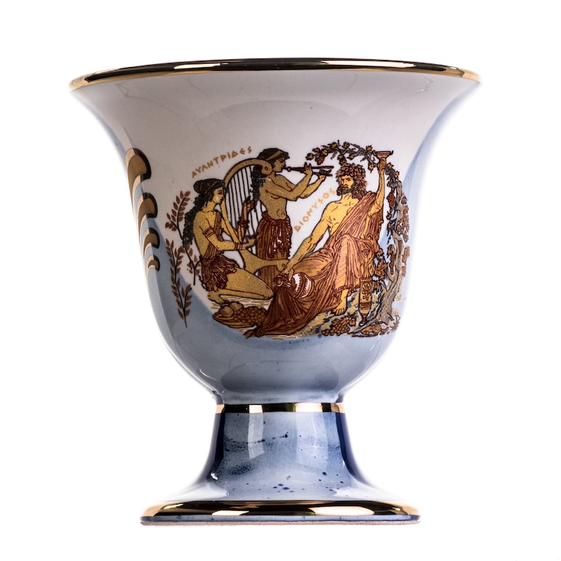 May include: A white ceramic goblet with a blue and gold design. The goblet features a scene of three figures, two women and a man, with the text "AYANTPIAS" and "DIOMYSOS" on the side.