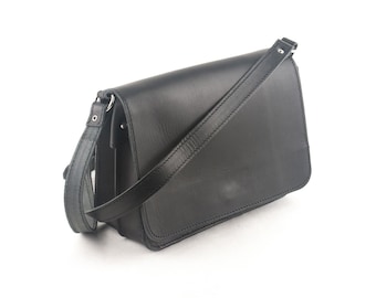 Op de afbeelding: Een zwarte leren messenger bag met een klepsluiting en verstelbare schouderriem. De tas is rechthoekig en heeft een gestikt ontwerp langs de randen. De riem is ongeveer 122 cm lang.