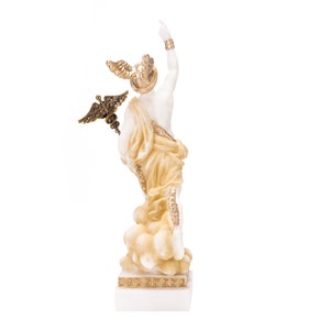 Puede incluir: Estatua de m&aacute;rmol blanco de una figura alada con detalles dorados. La figura est&aacute; de pie sobre una base en forma de nube y sostiene un bast&oacute;n con una serpiente enroscada alrededor. La estatua est&aacute; sobre una base de m&aacute;rmol blanco con un borde dorado.