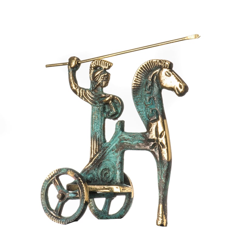 May include: Bronze-toned figurine depicting a chariot with a figure holding a spear, pulled by a horse. The chariot and figure have a verdigris patina, while the horse and spear are gold-colored. The wheels of the chariot are visible.