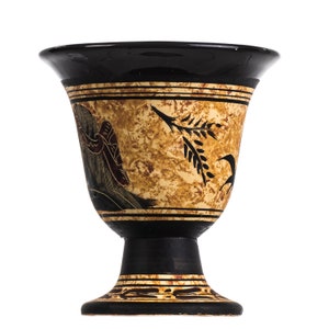 May include: A black and gold ceramic goblet with a stylized design of a figure and a plant. The goblet has a wide, flared rim and a tall, slender stem.
