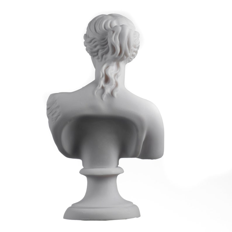 May include: White marble bust sculpture of a woman with her hair pulled back in a bun. The bust is on a white pedestal.