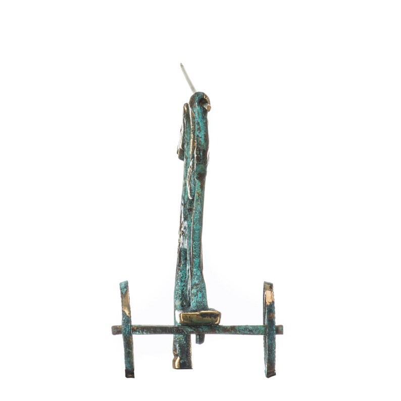 May include: A bronze-colored, miniature chariot with two wheels and a vertical structure. The chariot has a patina finish with hints of teal and gold. A small, silver-colored pin extends from the top.