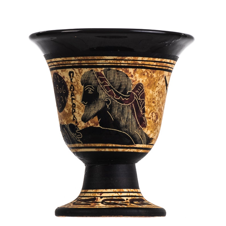 May include: Black and gold ceramic goblet with a black and white image of a man's head with a red and white headband. The goblet has a black base and a gold rim.