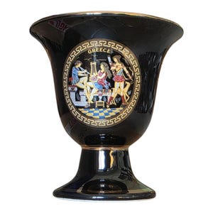 Peut inclure: Un calice en céramique noire orné d'un médaillon circulaire à bordure dorée avec un motif de grecque. L'illustration centrale représente trois personnages en tenue antique avec le mot GREECE inscrit en or sur un sol en damier.