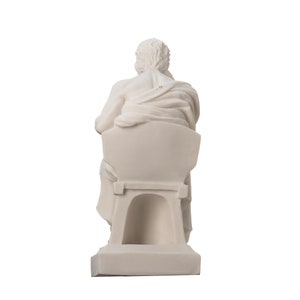 May include: White marble statue of a seated figure, the figure is facing away from the viewer, the statue is on a white base.