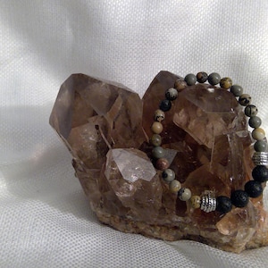 May include: A brown and gray gemstone bracelet with silver accents rests on a cluster of clear quartz crystals.