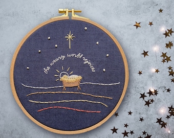 May include: Embroidered hoop art with a blue background and gold thread. The text reads "the weary world rejoices" with a star and a manger scene in the centre.