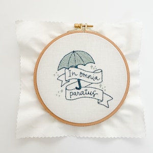 May include: Embroidered hoop art featuring a blue and green umbrella with the text "In omnia paratus" in a banner.