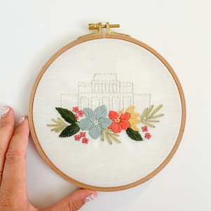 Puede incluir: Arte de bordado en aro con un edificio blanco con un diseño floral. Las flores son rosas, amarillas y azules con hojas verdes.