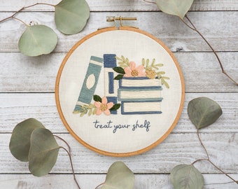 Könnte beinhalten: Stickrahmen-Kunst mit Stickerei, die einen Stapel blauer und weißer Bücher mit rosa Blumen und grünen Blättern zeigt. Der Text "treat your shelf" ist unter den Büchern gestickt.