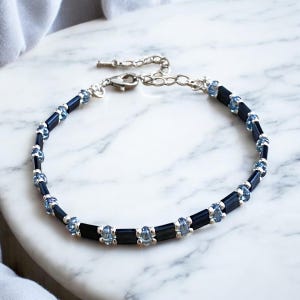 Peut inclure: Un bracelet délicat avec des perles noires et bleu clair alternées, entrecoupées de petits accents argentés. Le bracelet a une chaîne et un fermoir argentés pour un ajustement réglable. Le bijou est présenté sur une surface en marbre blanc.