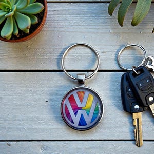 Peut inclure: Un porte-clés argenté avec le logo Volkswagen aux couleurs de l'arc-en-ciel. Le porte-clés rond est attaché à un anneau argenté. À côté du porte-clés se trouvent des clés de voiture avec des télécommandes noires et une clé argentée.