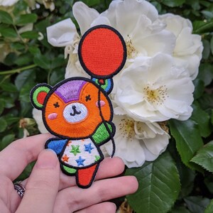 May include: An orange and purple embroidered patch of a cartoon bear holding a red balloon. The bear is wearing a blue, green and white star-patterned dress.
