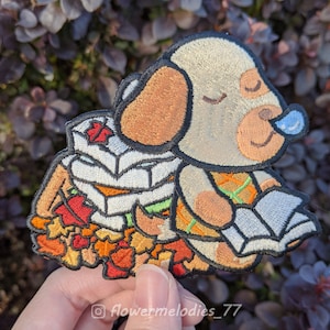 Peut inclure: Patch brodé représentant un chien blanc aux oreilles brunes portant une pile de feuilles blanches et orange. Le chien porte une chemise à motifs bruns et orange et a les yeux fermés. Le chien est entouré d'un tas de feuilles orange, rouges et jaunes.