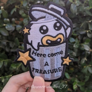 Może przedstawiać: Naszywka haftowana w kolorze niebieskim i białym z kreskówkowym duchem w koszu na śmieci. Duch ma na sobie czapkę i ma zaskoczony wyraz twarzy. Tekst "Here come a treasure" jest wyszyty na koszu na śmieci.