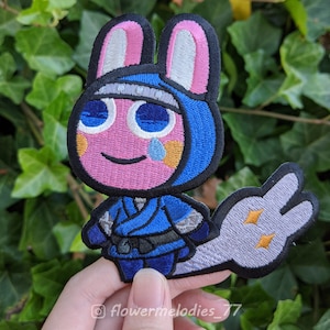 May include: A pink bunny rabbit wearing a blue ninja outfit with a white headband. The bunny has a tear coming from its eye and a white tail with two gold stars. The bunny is embroidered on a black fabric patch.