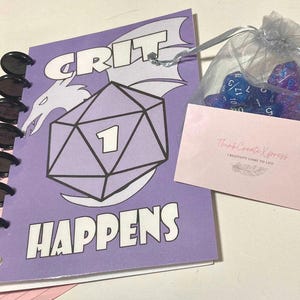 May include: Purple notebook with a white dragon and a large purple die with the number 1 on it. The notebook says "CRIT HAPPENS" in white letters. The notebook has a ring binder and is open to a blank page. There is a small bag of blue and purple dice in the background.