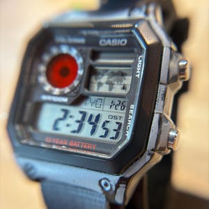 May include: Close-up of a Casio digital watch with a black strap. The watch face displays the time, date, and a world map icon. The watch has a red circular dial and the text "10 YEAR BATTERY". The watch is black and silver.