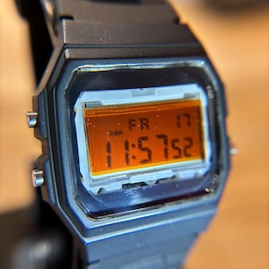 May include: Black digital watch displaying the time 11:57:52 on a rectangular orange screen. The watch band and case are black. The date is Friday the 17th. The watch has silver-colored buttons on the sides.