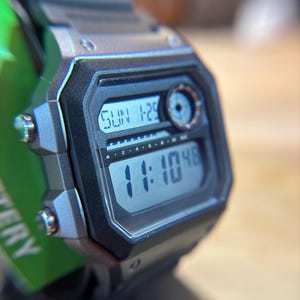 May include: A digital wristwatch with a black and silver case and a black strap. The watch face displays the day, date, and time. The watch is a rugged, sporty style.