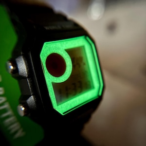 May include: Close-up of a black digital watch with a glowing green display. The watch face shows the time and a red circle. The watch is partially visible, with a green battery indicator on the side.