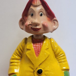 May include: A ceramic figurine of a gnome wearing a red hat and a yellow coat with green accents. The gnome has a large nose and a friendly smile.