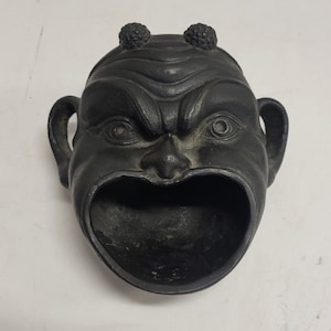 May include: A dark gray, cast metal ashtray in the shape of a stylized face with an open mouth. The face has a stern expression with furrowed brows, small ears, and two textured protrusions on top of the head.