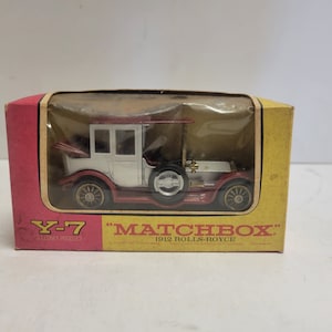 May include: A red and white toy car in a yellow and red box. The box says "Matchbox" and "1912 Rolls-Royce". The car is a replica of a vintage Rolls-Royce.