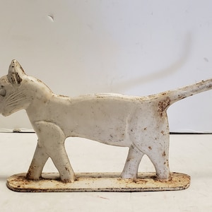 Puede incluir: Un tope de puerta de hierro fundido blanco en forma de gato con la cola levantada. El gato camina con la cabeza girada hacia un lado.
