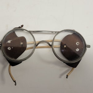 Könnte beinhalten: Eine vintage-Schutzbrille aus silberfarbenem Metall mit braunen Lederseitenschildern. Die Brille hat eine runde Form und ist durch eine Metallstange verbunden.