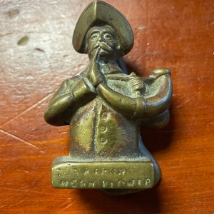 Pode incluir: Um batedor de porta de latão em forma de homem usando um chapéu e segurando um chifre. O batedor é inscrito com o texto "A.F. GROVER,  MFG. CO. NEW YORK."