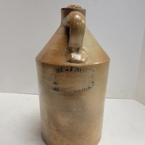 以下が含まれることがあります： ハンドルとコルク栓が付いた、ヴィンテージの炻器のジャグ。ジャグはタン色で、「UE-FRUIT」と「UNTAIN SYRUPS, NGERFORD, ROCHESTER, N.Y.」と書かれた黒い文字が書かれています。ジャグは円筒形の本体と円錐形のトップを持っています。
