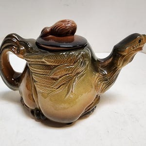 Puede incluir: Una tetera de cerámica con forma de camello con un esmalte marrón y verde. La tetera tiene una tapa con un pequeño pomo en la parte superior. La cabeza del camello está levantada y su cuello está curvado.