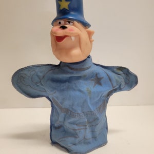 Puede incluir: Un títere azul y amarillo de un policía de dibujos animados con una cabeza grande y una sonrisa de dientes. El títere lleva un uniforme azul con una estrella en el pecho y un sombrero azul con una estrella amarilla.