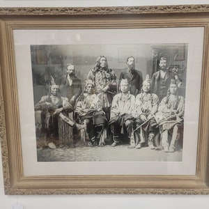 May include: A black and white photograph of a group of Native American men in traditional clothing. The men are seated in a row, and they are all looking at the camera. The photograph is framed in a wooden frame.