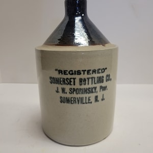 Puede incluir: Una jarra de cerámica blanca con la parte superior esmaltada en negro. La jarra tiene el texto "REGISTERED SOMERSET BOTTLING CO. J. W. SPORINSKY, PROP. SOMERVILLE, N. J." impreso en la parte delantera.