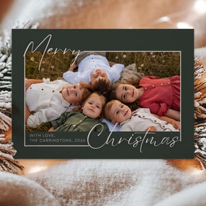 Puede incluir: Una tarjeta de felicitación navideña rectangular con un borde verde oscuro que presenta una fotografía central de cinco niños acostados en círculo sobre el césped. La tarjeta incluye el texto cursivo blanco Merry Christmas y With Love, The Carringtons, 2026.
