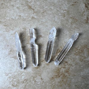 May include: Four clear quartz crystal points of varying sizes are displayed on a neutral-toned surface. The crystals are translucent, with visible internal structures and sharp, defined edges. They are arranged in a slightly scattered pattern.