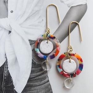 Puede incluir: Un par de pendientes de aro dorados con detalles redondos de resina multicolores y superpuestos y un círculo blanco moteado en el centro de cada detalle.