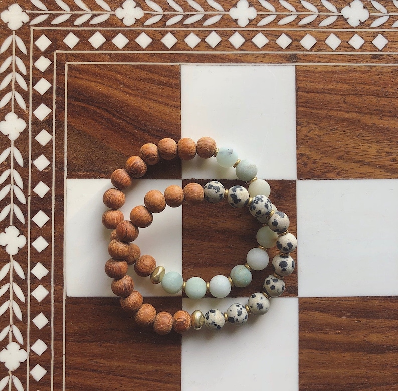 May include: Two beaded bracelets stacked on top of each other. The bottom bracelet is made of brown wood beads. The top bracelet is made of white and black speckled beads and light green beads.
