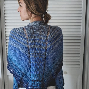 May include: A blue and gray knitted shawl with a central cable design. The shawl features a gradient of blues, from light to dark, and a scalloped edge. The back of the shawl is visible, showcasing the intricate knit pattern.
