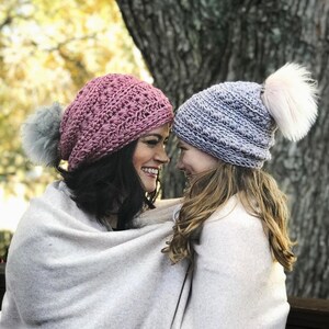 Può includere: Due persone che indossano cappelli all'uncinetto. La persona a sinistra indossa un cappello rosa con un pon pon bianco e la persona a destra indossa un cappello lilla chiaro con un pon pon bianco.