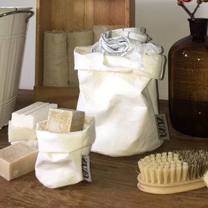 Könnte beinhalten: Eine Holzkiste mit drei weißen Handtüchern, eine weiße Stofftasche mit dem Aufdruck "ZUZA", eine braune Glasflasche mit einem Zweig mit roten Beeren und zwei Holzbürsten mit Naturborsten.