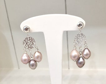 May include: Elegant silver chandelier earrings with three teardrop-shaped pearls in shades of pink and gray. The earrings feature a delicate filigree design and are displayed on a white jewelry stand.