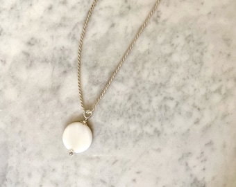 May include: A silver necklace with a white, circular pendant. The pendant is attached to a delicate silver chain. The necklace is displayed on a marble surface. The necklace is a piece of jewelry.
